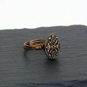 May include: A copper wire ring with a black and gold circular design. The design features a stylized floral pattern.