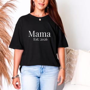 Puede incluir: Camiseta negra con la palabra "Mama" en blanco, con "Est: 2026" debajo. La camiseta se usa con vaqueros azul claro. La imagen muestra a una persona usando la camiseta.