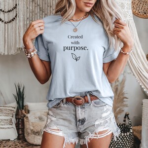 Puede incluir: Camiseta azul claro con mangas enrolladas, con el texto "Created with purpose." en escritura negra, acompañada de un diseño de hojas. La camiseta se combina con pantalones cortos de mezclilla y un cinturón marrón.