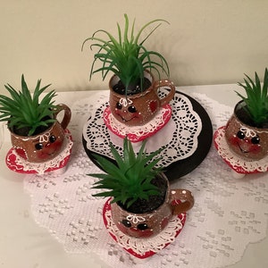 May include: Four small brown ceramic pots with painted gingerbread faces, each holding a green succulent plant. The pots are on white doilies with red trim and are arranged on a black and white doily.