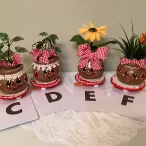 May include: Four small brown ceramic pots with painted faces, each with a different plant or flower. The pots are decorated with red and white gingham ribbon, white crochet trim, and red hearts. The pots are sitting on small red and white plates, and each pot is labeled with a letter: C, D, E, and F.