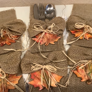 May include: Six burlap place card holders with a fall theme. Each holder features a brown burlap bow, a brown and orange leaf, and a small piece of straw.