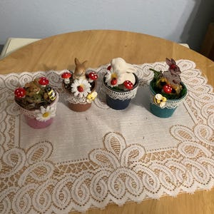 May include: Four small decorative pots with bunny figurines, mushrooms, and flowers. The pots are painted in pink, brown, blue, and teal, and are decorated with lace trim. A bee and ladybugs are also included.