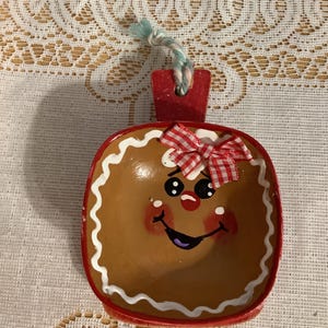 May include: A hand-painted, gingerbread-themed ornament. The ornament is brown with a red border and a white icing design. It features a smiling face, a red bow, and a blue and white string for hanging. A festive holiday decoration.
