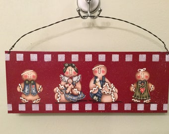 May include: A rectangular red wooden sign featuring four painted gingerbread figures. The sign has a white checkered border and is suspended by a dark wire. The figures include a gingerbread person in a vest, one holding a smaller cookie, one decorating a heart, and one in a dress.