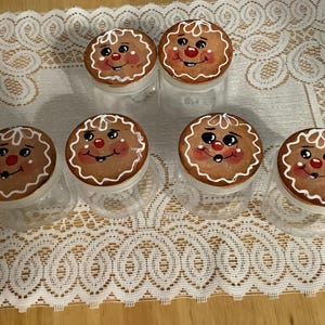 May include: Six clear glass jars with gingerbread man lids. The lids are round, brown, and decorated with white icing details, red noses, and smiling faces. The jars are arranged on a white lace tablecloth.
