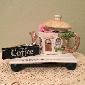 May include: A white ceramic teapot shaped like a cottage with a thatched roof, filled with pink tea bags. The teapot is on a white wooden stand with black legs and a sign that says "Coffee".