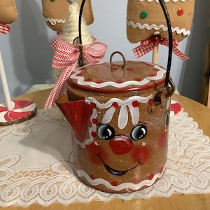 May include: A brown, gingerbread-themed metal teapot with a lid and handle. The teapot is decorated with a smiling gingerbread man face, white frosting details, and red accents. The teapot is sitting on a lace tablecloth.