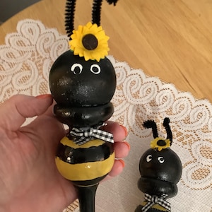 May include: Two wooden bee figurines. The bees are painted black and yellow with a sunflower on their heads and a black and white checkered bow. One bee is held in a hand. The background is a wooden table with a white lace doily.