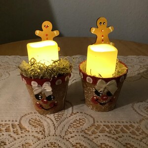 May include: Two decorative candle holders with a gingerbread man theme. Each holder is a small, brown pot with a painted face, a red rim, and a white bow. A yellow, rectangular candle sits inside, topped with decorative straw, and a wooden gingerbread man.