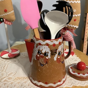 May include: A brown metal pitcher with a gingerbread man design, holding kitchen utensils. The pitcher has a red handle and spout, and a matching lid with a red ball on top. Decorative gingerbread man ornaments are in the background.