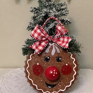 May include: A gingerbread man ornament with a red and white checkered bow, a red nose, and a smiling face. The ornament is hanging from a small artificial Christmas tree.