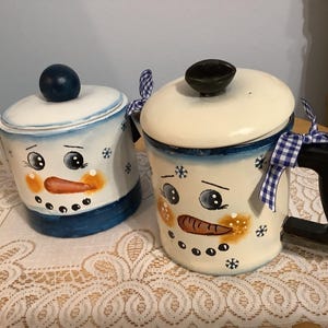 May include: Two snowman-themed ceramic containers. One is a teapot with a blue handle and lid knob. The other is a mug with a black handle. Both have painted snowman faces, with carrot noses and coal eyes.