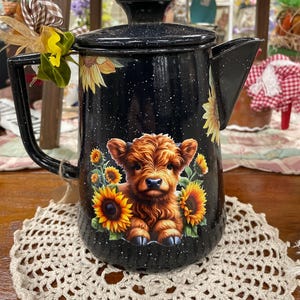 May include: Black enamel teapot with a cow and sunflower design. The teapot has a black handle and spout. The cow is brown and surrounded by yellow sunflowers. The teapot is sitting on a cream-colored, crocheted doily.