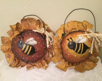 May include: Two handmade bee and sunflower decorations. Each decoration is a round, brown and yellow painted canvas with a black and yellow bee painted on it. The canvas is surrounded by a yellow fabric that resembles sunflower petals. Each decoration has a black wire hanger.