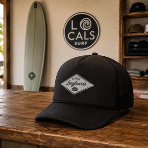 May include: A black trucker hat featuring a white diamond-shaped patch on the front. The patch displays the text Classic Longboards Est. 1979 in a black script and sans-serif font. The hat has a solid front panel and a mesh back panel.