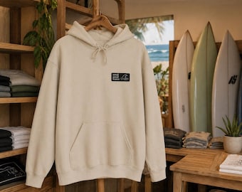 Könnte beinhalten: Cremefarbener Kapuzenpullover mit Fronttasche und Kordelzugkapuze. Ein kleines schwarzes rechteckiges Patch auf der Brust zeigt "send good waves" mit einer Wellengrafik. Der Hoodie hängt an einem Holzregal, mit Surfbrettern im Hintergrund.