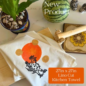 May include: A white 27in x 27in kitchen towel with a linocut design. The towel features an orange sun, gold circles, and a black and white chicken illustration. The words "New Product" are visible.