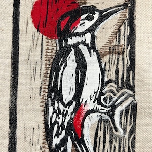 May include: A black, white, and red woodpecker illustration on a beige background. The bird is perched on a tree branch, with a red sun in the background. The image is framed in a black border.