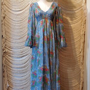 May include: A vintage blue maxi dress with a floral pattern, long sleeves, and a V-neck. The dress is displayed on a mannequin with a brown afro wig. The dress has a flowing silhouette and bell sleeves.