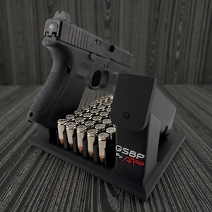 Könnte beinhalten: Eine schwarze Handfeuerwaffe liegt auf einem schwarzen Ständer, zusammen mit einem Magazin und einer Munitionsschale. Der Ständer trägt den roten Schriftzug "GSBP by AP". Der Hintergrund ist ein dunkles Holzmaserungsmuster.