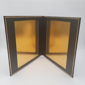 May include: A dark brown diploma or certificate folder featuring two interior panels. Each panel has a central rectangular gold foil insert framed by a thin gold border, providing a reflective surface for displaying documents or awards.