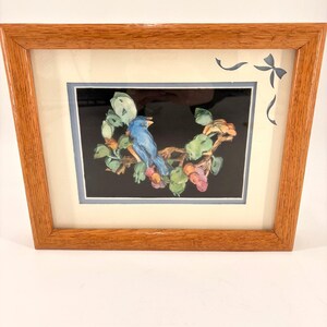 May include: A framed picture of a blue bird on a branch with green leaves and red berries, set against a black backdrop. The artwork is in a light brown wooden frame, with a cream-coloured mount and a blue bow design in the top right corner.
