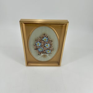 Op de afbeelding: Een ingelijste bloemschikking met blauwe bloemen met gele centra en bruine accenten, tegen een lichtgroene ovale achtergrond. De schikking is vastgebonden met een wit lint en gevat in een goudkleurige lijst.