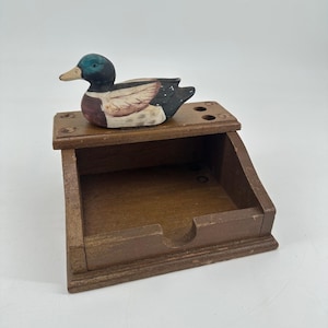 May include: A wooden duck-shaped desk organizer. The duck figurine is painted with a green head, brown body, and white and black wings. The organizer has a drawer and several holes on top for pens or pencils.