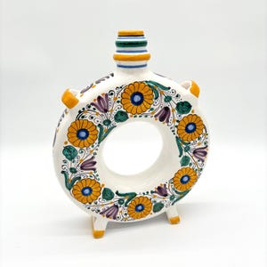 May include: A white ceramic donut-shaped vase featuring hand-painted floral motifs in yellow, purple, and green. The vessel has a central circular opening, two side handles, a narrow neck with blue and yellow bands, and two small feet.
