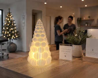 Op de afbeelding: Een witte, kegelvormige lamp in de vorm van een kerstboom, bestaande uit meerdere gestapelde, afgeronde, schijfvormige segmenten. De structuur heeft een getextureerd, geribbeld uiterlijk en verspreidt een warme, zachte gloed van binnenuit.