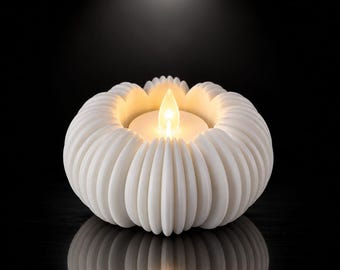 Op de afbeelding: Een witte, ronde kaarshouder met een geribbeld, bloembladachtig ontwerp aan de buitenkant. De houder bevat een centraal, gloeiend LED-theelichtje dat een warm, zachtgeel licht uitstraalt en een contrast vormt met de donkere achtergrond.