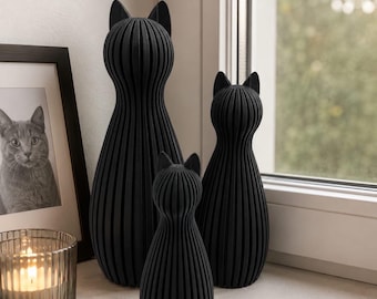 Op de afbeelding: Drie zwarte kattenbeeldjes met een verticale geribbelde textuur. De beelden hebben een ronde kop met puntige oren en een taps toelopend, langwerpig lichaam. De set bevat een grote, een middelgrote en een kleine rechtopstaande kat.