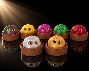 Op de afbeelding: Zeven ronde, knoedelvormige figuren met grote, expressieve ogen, elk rustend in een klein bruin mandje met vlechtwerk. De figuren zijn paars, geel, magenta, groen, rood, zilver en goudkleurig en zijn in twee rijen tegen een zwarte achtergrond geplaatst.