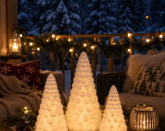 Op de afbeelding: Drie conische, witte, getextureerde decoratieve bomen met een gloeiend, gebobbeld oppervlak. Deze verlichte ornamenten zijn gerangschikt op een houten oppervlak samen met een kleine glazen kaarsenhouder, dennenappels en dunne lichtsnoeren.