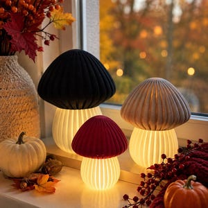Könnte beinhalten: Drei pilzförmige Lampen mit gerippten Kappen in Schwarz, Rot und Beige. Jede Lampe verfügt über einen weißen, vertikal geschlitzten Sockel, der ein warmes, leuchtendes Licht abgibt. Die Lampen sind zusammen angeordnet und beleuchten die Fläche.