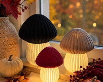 Op de afbeelding: Drie paddestoelvormige lampen met geribbelde kappen in zwart, rood en beige. Elke lamp heeft een witte, verticaal gespleten voet die een warm, gloeiend licht uitstraalt. De lampen staan bij elkaar en verlichten het oppervlak zachtjes.