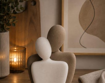 Op de afbeelding: Een decoratief beeldhouwwerk met twee abstracte menselijke figuren in een omhelzing. De voorste figuur is glad en wit, terwijl de figuur erachter getextureerd en bruin is. Beide figuren zijn gezichtsloos en hebben afgeronde vormen.