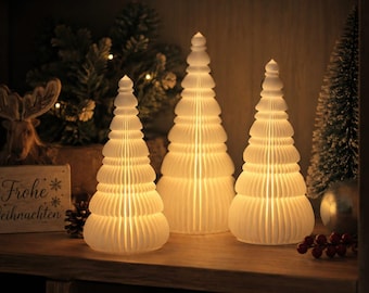 Op de afbeelding: Drie witte, gelaagde, kegelvormige papieren decoraties in de vorm van kerstbomen, van binnenuit verlicht door een warm licht. De structuren hebben een geplooid, accordeonachtig ontwerp met een puntige bovenkant.