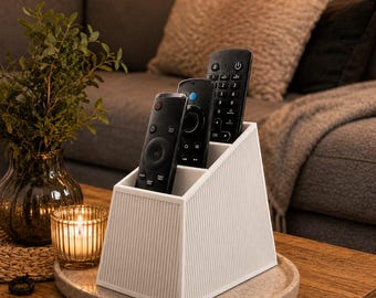 May include: A white, trapezoidal remote control holder with a vertical ribbed texture on the exterior. The organizer features three distinct compartments of varying heights, currently holding three black television remote controls.