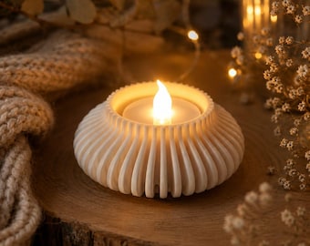 Op de afbeelding: Een witte, ronde kaarshouder met een geribbeld, verticaal lattenontwerp. De houder heeft een centrale opening met een klein, op batterijen werkend LED-theelichtje met een vlamvormig lampje dat een zacht, goudkleurig licht uitstraalt.