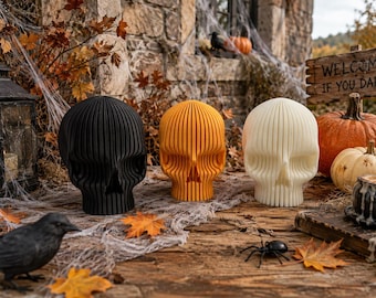 Op de afbeelding: Drie schedelsculpturen op een rij, bestaande uit een zwarte, een oranje en een gebroken witte schedel. Elke schedel is opgebouwd uit een reeks verticale, parallelle lamellen die een geribbelde, gelaagde uitstraling geven aan de hele structuur.