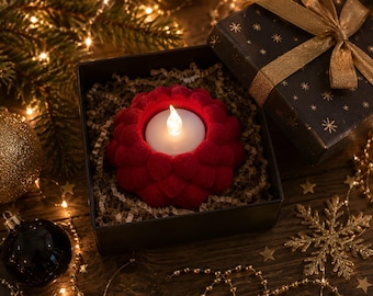 Op de afbeelding: Een rode, getextureerde, bloemvormige kaarshouder met een wit LED-theelichtje. De kaarshouder zit in een zwarte geschenkdoos gevuld met papierversnippering, naast gouden en zwarte kerstversiering en lichtjes.
