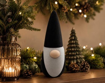 Op de afbeelding: Een decoratief kabouterbeeldje met een hoge, puntige zwarte muts, een lichtgrijze baard en een ronde beige neus. De kabouter heeft een glad, afgerond lichaam en staat rechtop, wat een strak ontwerp met een duidelijk kleurcontrast laat zien.