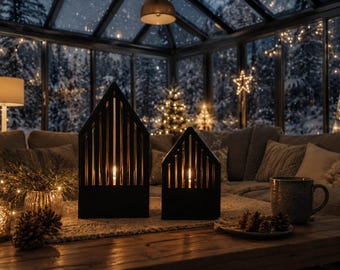 Op de afbeelding: Twee zwarte kaarsenhouders in de vorm van een huis met verticale latten, elk met een brandende kaars. De structuren hebben een puntdak en stralen een warme gloed uit, wat een contrast vormt met de donkere, minimalistische buitenframes.