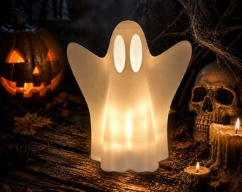 Op de afbeelding: Een witte, doorschijnende, spookvormige lamp met twee ovale ooguitsparingen. De lamp straalt van binnenuit een warme, zachte gloed uit die de gladde, ronde vorm verlicht. De figuur heeft uitgestrekte armen en een golvend basisontwerp.