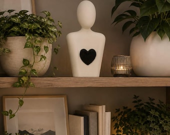 Op de afbeelding: Een gladde, gebroken witte abstracte menselijke buste sculptuur met een opvallend, getextureerd zwart hart in het midden van de borst. Het figuur heeft een ovaalvormig hoofd zonder gelaatstrekken en een torso die naar de basis toe versmalt.