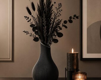 May include: A matte black ribbed vase filled with a bouquet of dried black pampas grass, eucalyptus, and various stems. Beside the vase are two candles, one tall and textured, the other shorter and ribbed, both emitting a warm, glowing flame.