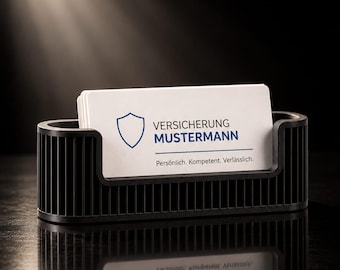 Op de afbeelding: Een zwarte, geribbelde visitekaarthouder met een stapel witte kaarten. De bovenste kaart bevat een blauw schildicoon, de tekst "VERSICHERUNG MUSTERMANN" en de slogan "Persönlich. Kompetent. Verlässlich." in zwarte letters.