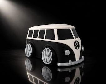 Op de afbeelding: Een zwart-witte speelgoedbus met het Volkswagen-logo op de voorzijde en op elk van de vier zilverkleurige wielen. Het voertuig heeft een wit dak en voorpaneel, zwarte zijpanelen met rechthoekige ramen en een zilverkleurige voorbumper.